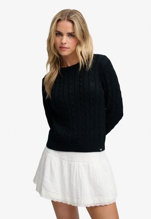 Lyshåret kvinde iført en sort kabelstrikket sweater og et hvidt nederdel med blondekant, stående mod en ensfarvet baggrund.