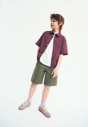 Junge trägt ein kurzärmliges lila Hemd über einem weißen T-Shirt, grüne Shorts, graue Socken und beige Turnschuhe, steht mit den Händen in den Taschen.