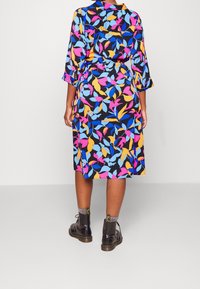 Kvinna i en färgglad midi-klänning med lövmönster och trekvartslång ärm, stående mot en enhetlig bakgrund.