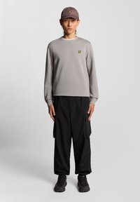 Szürke pulóver kerek nyakkivágással és sárga logóval a mellkasán, fekete cargo nadrággal és sötét sapkával párosítva. Texturált tornacipők egészítik ki a megjelenést.