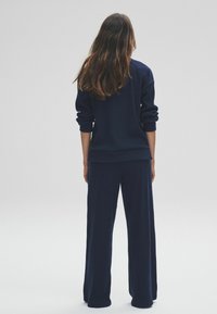 Femme aux longs cheveux bruns portant un pull côtelé bleu foncé et un pantalon large, se tenant de dos contre un fond gris uni.