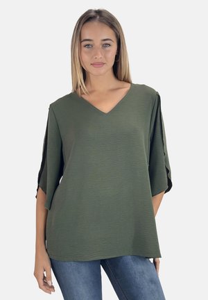 Top verde oliva a scollo a V con maniche ampie, tessuto strutturato e un sottile motivo a onde. Indossato con jeans blu, presenta aperture laterali.