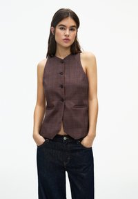 Brun rutig ärmlös väst med hög hals, som har fem svarta knappar och framfickor, tillsammans med mörkblå denimjeans.