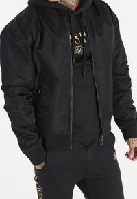 Giubbotto bomber nero realizzato in tessuto lucido, dotato di chiusura con zip, polsini a coste e logo ricamato sul petto, abbinato a un hoodie nero.