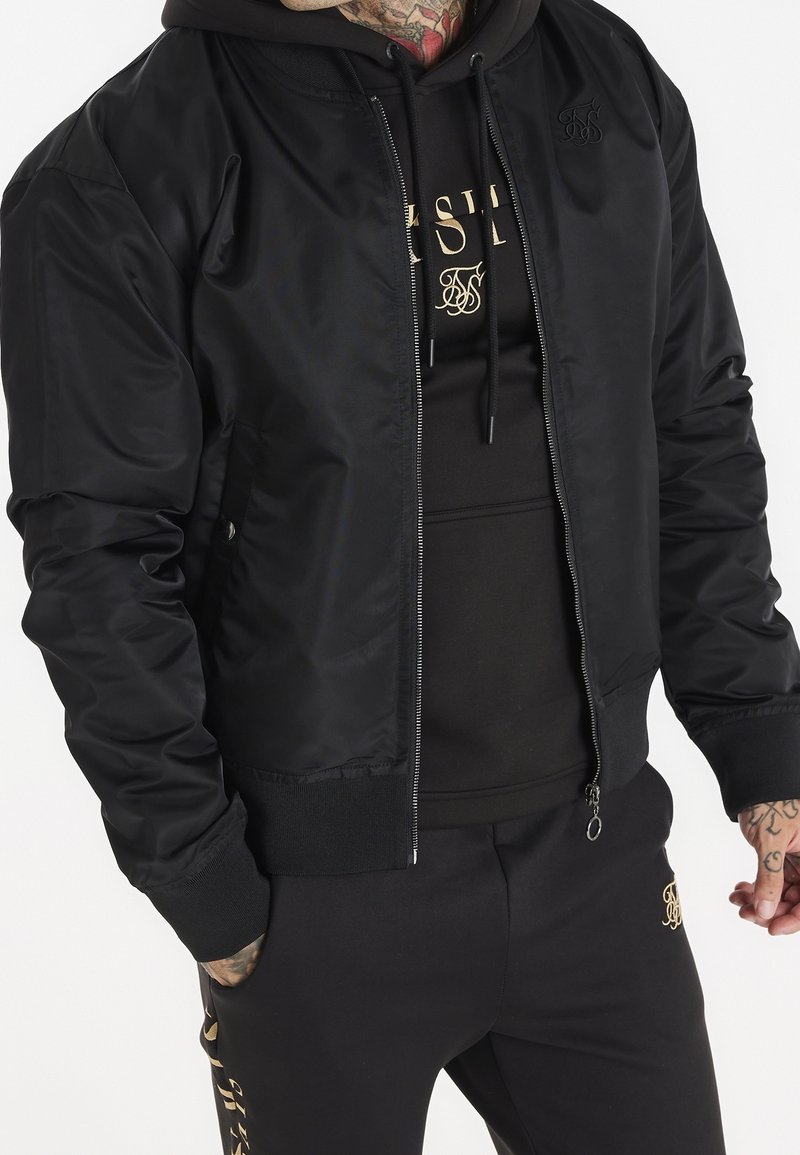 Giubbotto bomber nero realizzato in tessuto lucido, dotato di chiusura con zip, polsini a coste e logo ricamato sul petto, abbinato a un hoodie nero.