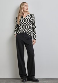 Zwart-wit geblokte blouse met geometrische ontwerpen, gecombineerd met hoge zwarte broek. Het model staat op een gestructureerd grijs oppervlak.