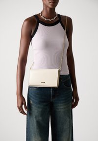 Borsa a tracolla in pelle crema con una tracolla in catena dorata, dalla finitura liscia e dalla forma rettangolare piccola. Indossata sopra una canotta lavanda e jeans.
