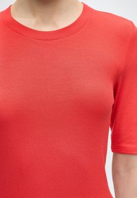 Κοντινό πλάνο ενός ατόμου που φοράει πλεκτό πουκάμισο κοραλλί με ριμπ ύφανση, κοντά μανίκια και στρογγυλή λαιμόκοψη, μπροστά σε απλό φόντο.