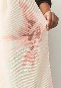 Audums ar mīkstu, ribotu tekstūru baltā krāsā ar bālām rozā un brūnām abstraktām rakstām, pārklāts un turēts ar roku.