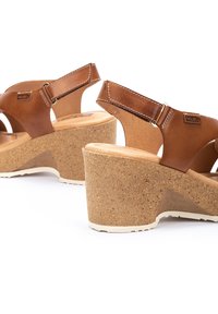 Sandalias de cuña de cuero marrón con plataforma de corcho. Cuenta con una correa ajustable en el tobillo y un diseño de punta abierta. Detalle de costura en las correas.