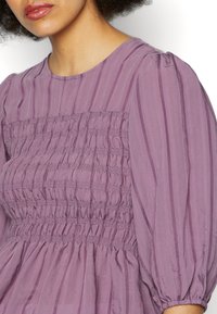 Femme portant un chemisier violet à manches longues avec des détails froncés et un tissu rayé verticalement.