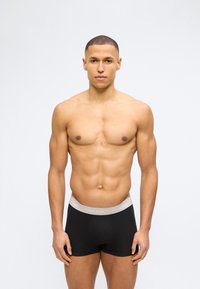 Άνδρας χωρίς μπλούζα, κοιτάζοντας προς τα εμπρός, φοράει μαύρα μποξέρ Calvin Klein με γκρι μέση, πάνω σε απλό φωτεινό φόντο.