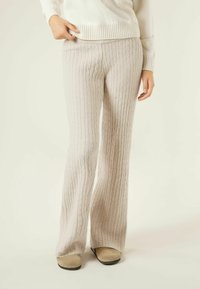 Beżowe spodnie o szerokich nogawkach z dzianiny warkoczowej z prążkowaną fakturą, miękkim wykończeniem i elastycznym paskiem w talii. W połączeniu z jasnym obuwiem.