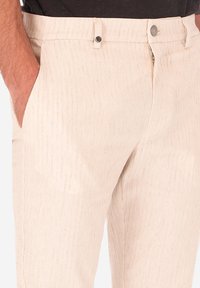 Pantaloni beige chiaro realizzati in un tessuto testurizzato con righe verticali, dotati di tasche inclinate e chiusura con bottone in vita.