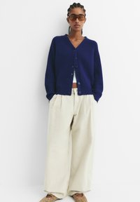 Mornarska rebrasta kardigan s V-izrezom i kopčanjem na gumb, uparen s labavim bež hlačama i uzorkovanim cipelama na klizanje.