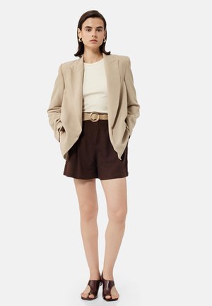 Kvinde iført beige blazer, hvid top, mørkebrune shorts med flettet bælte, guld øreringe i form af hoops og mørkebrune åbne hæle, stående mod en hvid baggrund.