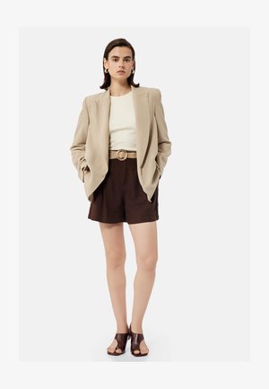 Kvinde iført beige blazer, hvid top, mørkebrune shorts med flettet bælte, guld øreringe i form af hoops og mørkebrune åbne hæle, stående mod en hvid baggrund.
