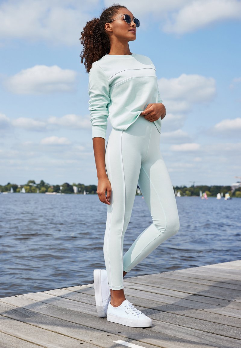 Lichtgroene sweatshirt met ronde hals en naaddetails, gecombineerd met groene leggings. Witte sneakers maken de sportieve look compleet. Achtergrond: water.