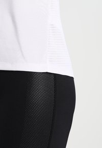 White athletic top with textured fabric, paired with black leggings featuring a mesh panel design. Smooth, fitted silhouette with contrasting colors.