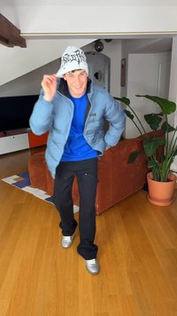 Svijetloplava jakna, kraljevsko plava košulja, crne hlače i srebrne cipele. Bijela kapa s crnim grafičkim uzorkom. Unutarnji prostor s biljkama.