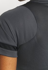 La camiseta atlética de color gris oscuro presenta una tela suave con acentos de malla, mangas cortas y un diseño ajustado para una mayor transpirabilidad.