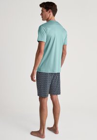 Lichtblauwe katoenen T-shirt, korte mouwen, gecombineerd met marineblauwe shorts met een wit abstract patroon. Model staat op blote voeten, achterkant zichtbaar.