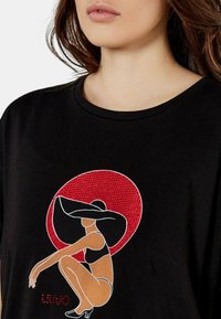 Maglietta nera in cotone con una grafica stilizzata in rosso e bianco di una figura con un cappello a tesa larga, su uno sfondo circolare rosso.