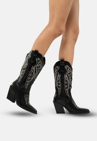 Botas de vaquero de cuero negro con diseños bordados en plata, punta afilada y un tacón de bloque texturizado, con tiradores y cierre de cremallera.