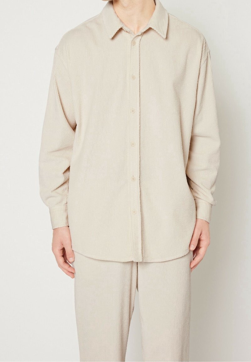 Chemise en velours côtelé beige à boutons, avec col, manches longues et coupe décontractée ; présente une texture côtelée verticale et des boutons à l'avant.