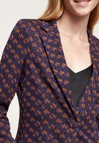 Lila kavaj med ett geometriskt mönster i orange och svart. Har en spetsig krage, en enda knapp och strukturerad tyg.