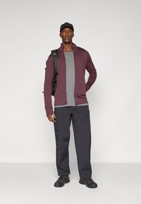 Jachetă zip-up maron peste un tricou gri, asociată cu pantaloni cargo gri închis și adidași negri, completată de o șapcă neagră și un rucsac.