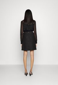 Vestido negro con parte superior transparente de manga larga con lunares y falda texturizada acampanada. Se lleva con tacones negros de tiras, mostrado desde atrás.