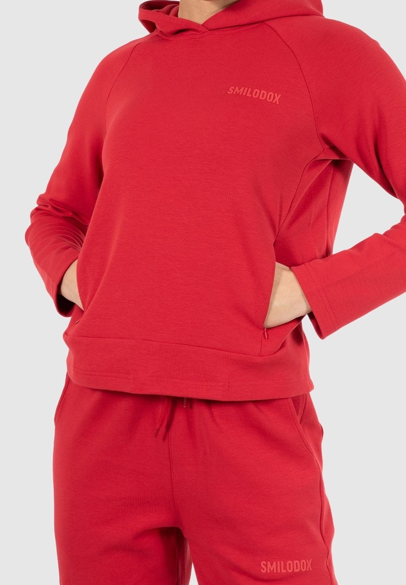 Hoodie rouge avec une poche kangourou à l'avant, coupe confortable et poignets côtelés. Le nom de la marque 'SMILODOX' est subtilement imprimé sur la poitrine.