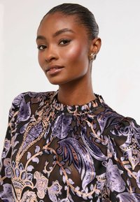 Caurspīdīga melna blūze ar purpura un zelta ziedu rakstiem, savilkti kakla apmale un pūkaini piedurknes. Modelis valkā zelta spirālveida auskari.