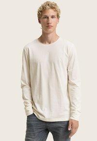 Junger Mann mit lockigem, blonden Haar, der ein schlichtes, cremefarbenes Langarmshirt und dunkelgraue Jeans trägt und vor einem beigen Hintergrund steht.
