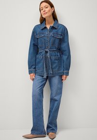 Giubbotto in denim con vita a cintura, tasche sul petto e maniche lunghe, abbinato a jeans svasati coordinati e scarpe beige a punta.