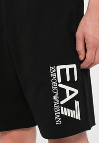 Black shorts made of lightweight fabric featuring a large white embroidered logo "EA7 Emporio Armani" on the left leg.