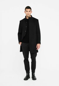 Cappotto nero con colletto alto, chiusura a bottoni sul davanti e tasche laterali. Abbinato a un maglione nero a trama e pantaloni neri aderenti, completati da stivali neri.