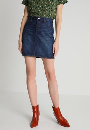 Persoon die een donkerblauwe denimrok, een groen shirt met luipaardprint en roodbruine puntige enkellaarsjes draagt, staand tegen een eentonige achtergrond.