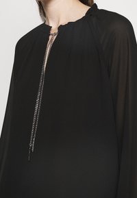 Schwarze Bluse mit hohem Kragen und Falten, einem Frontverschluss und einem filigranen Kettendetail. Der Stoff hat eine glatte, durchscheinende Textur.