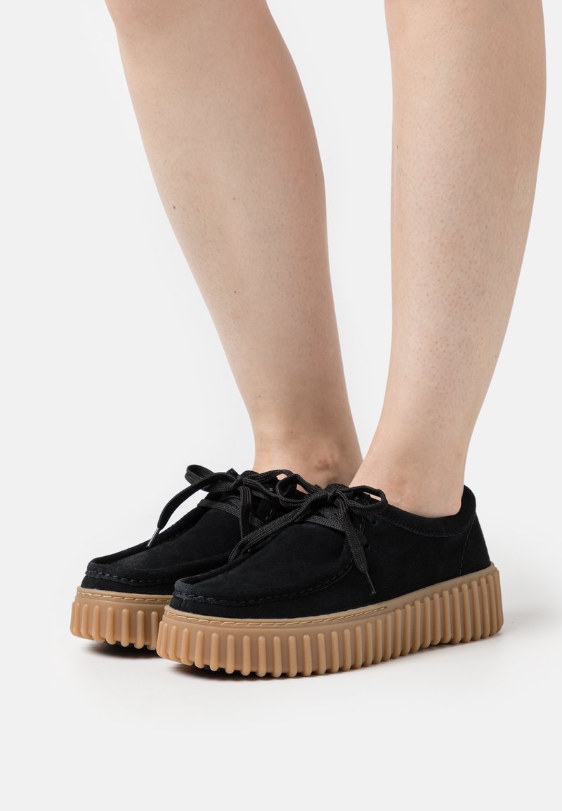Personne portant des chaussures en daim noir à lacets avec des semelles épaisses en caoutchouc rainurées, montrée de mi-mollet vers le bas sur un fond blanc.