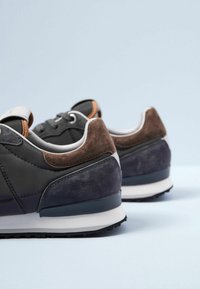 Ett par mörka sneakers med bruna mockadetaljer, vita mellansulor och svarta yttersulor på en ljusblå yta och bakgrund.