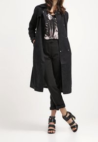 Kobieta ubrana w czarny długi płaszcz, wzorzysty top, czarne podwinięte spodnie i czarne sandały na wysokim obcasie, stojąca ze skrzyżowanymi nogami.