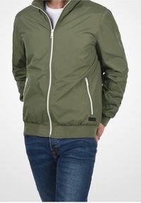 Chaqueta bomber verde oliva con cremallera frontal, detalles en blanco, dos bolsillos con cremallera y puños y dobladillo acanalados. Material liso y ligero.