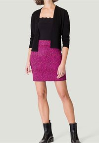 Cardigan noir à manches longues associé à une mini-jupe rose à motif éparpillé. Le mannequin porte des bottines noires brillantes.