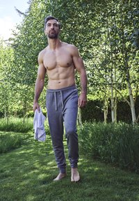 Grå sweatpants med elastisk talje, tætsiddende ved anklerne. En barbrystet mand går på græsset, mens han holder en hvid skjorte i hånden.