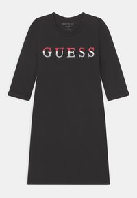 Robe noire à manches trois quarts, col rond, mettant en avant un logo "GUESS" audacieux en rouge et blanc sur le devant. Texture de tissu douce.