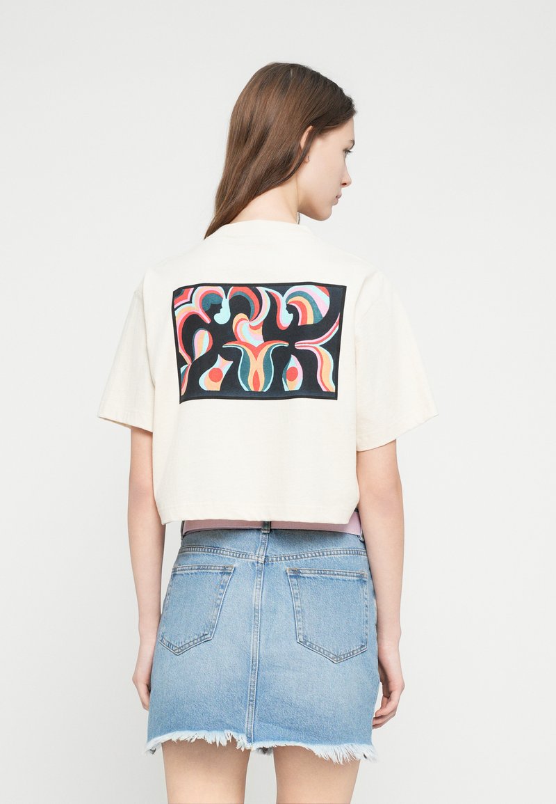 Κρεμ κοντομάνικη μπλούζα με πολύχρωμο αφηρημένο σχέδιο στην πλάτη. Συνδυάζεται με μίνι φούστα από ανοιχτό τζιν με ξεφτισμένο τελείωμα.