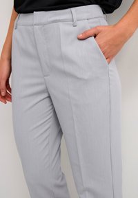 Ljusgrå skräddarsydda byxor med en slät textur, med framfickor och en slim fit-design, delvis sedd med en hand vilande i en ficka.