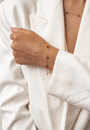Wrist wearing delicate gold bracelet with heart charms, paired with matching necklace, visible over white blazer sleeve.